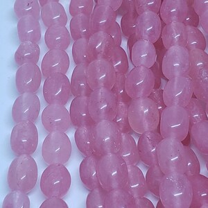 May include: Close-up of strands of polished, oval-shaped rose quartz beads. The beads are a soft pink color and have a smooth, glossy surface. The beads are strung together, creating a flowing, textured appearance. Ideal for jewelry making.
