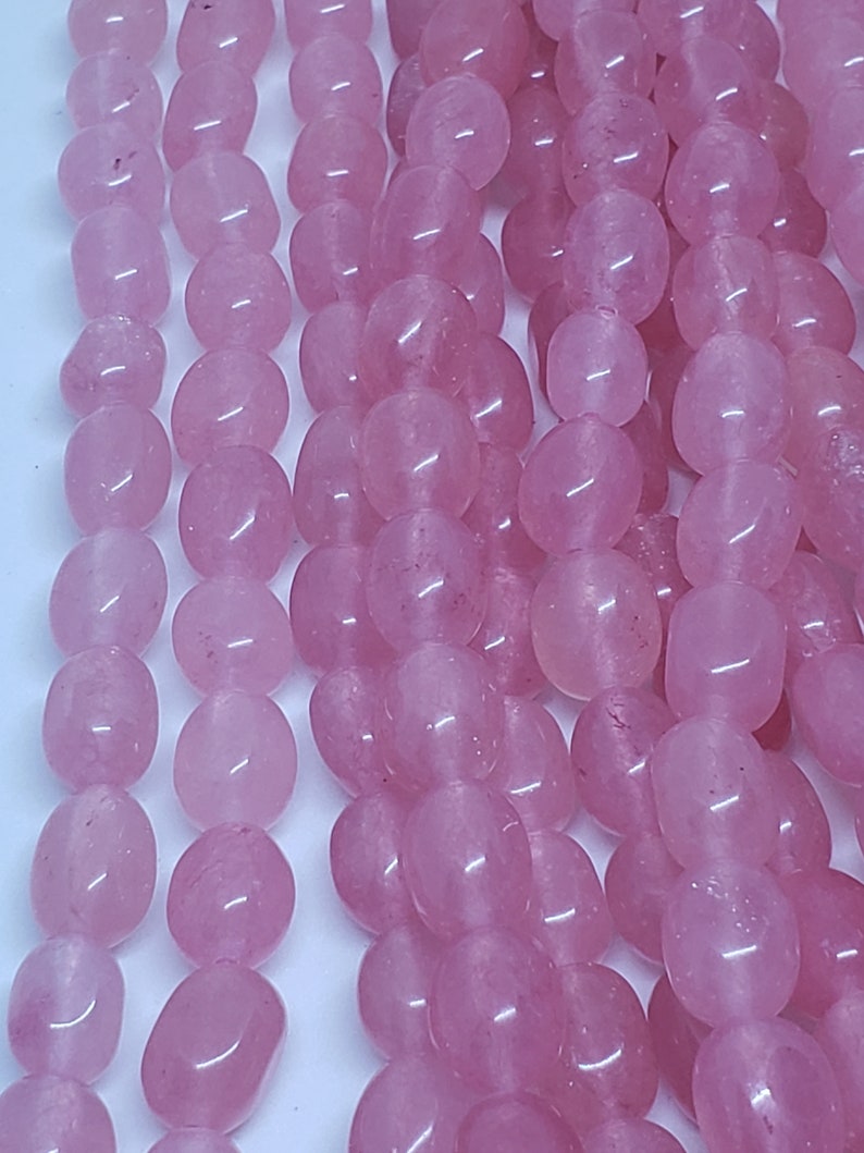 May include: Close-up of strands of polished, oval-shaped rose quartz beads. The beads are a soft pink color and have a smooth, glossy surface. The beads are strung together, creating a flowing, textured appearance. Ideal for jewelry making.