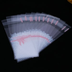 May include: A stack of clear plastic bags with a white lace and pink ribbon design. The bags are ideal for packaging small gifts or favours.