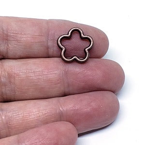 May include: A small, copper-colored flower-shaped charm. The charm is held in a hand against a white background. The charm has a five-petal design with a hollow center.