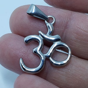 May include: A silver Om symbol pendant with a loop for a chain. The pendant is made of metal and has a shiny finish.