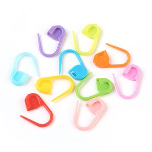 May include: A set of 12 colorful plastic stitch markers in various colors, including purple, yellow, green, blue, orange, pink, and red. The stitch markers are shaped like a loop with a small button on one end.
