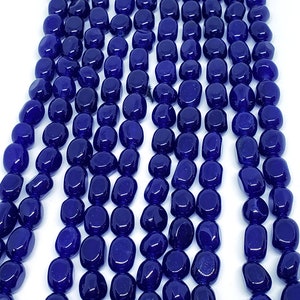 May include: Close-up of strands of polished, deep blue gemstone beads. The beads are oval-shaped and have a glossy finish. The beads are strung together, creating a flowing, textured pattern. The background is plain white, highlighting the color and texture of the beads.