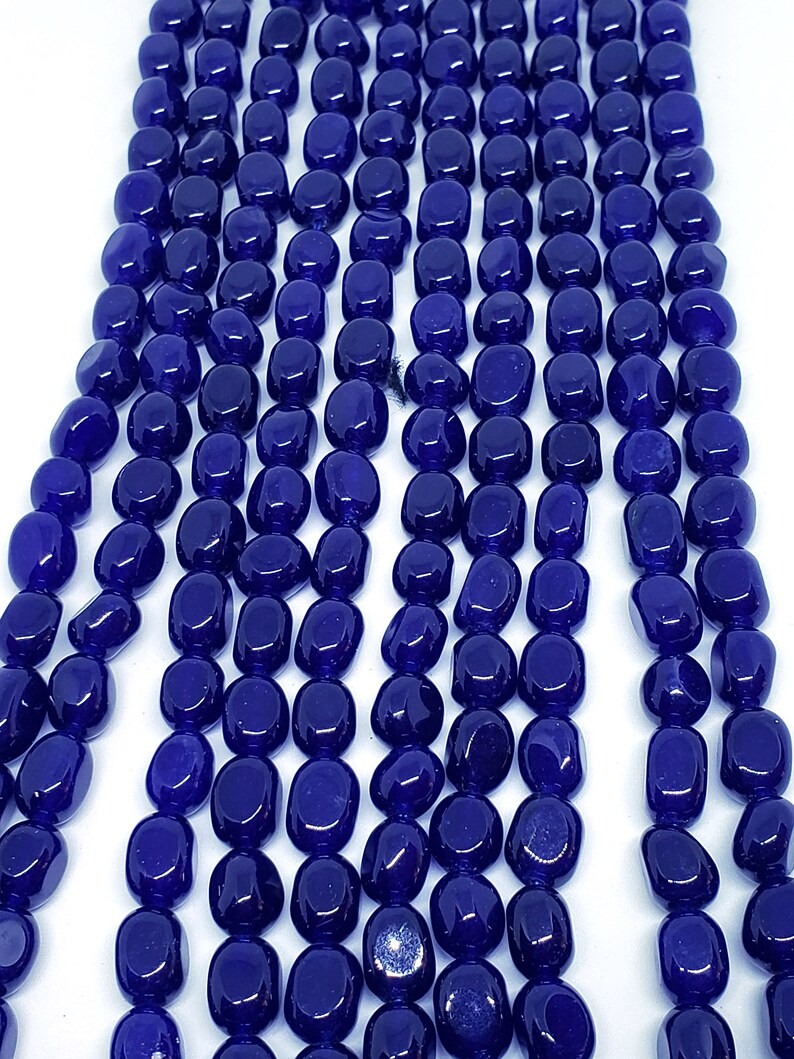 May include: Close-up of strands of polished, deep blue gemstone beads. The beads are oval-shaped and have a glossy finish. The beads are strung together, creating a flowing, textured pattern. The background is plain white, highlighting the color and texture of the beads.