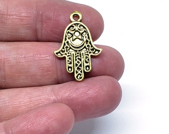Puede incluir: Encanto de metal dorado en forma de mano de hamsa con detalles intrincados. La mano de hamsa tiene un diseño de estrella y luna creciente en el centro.