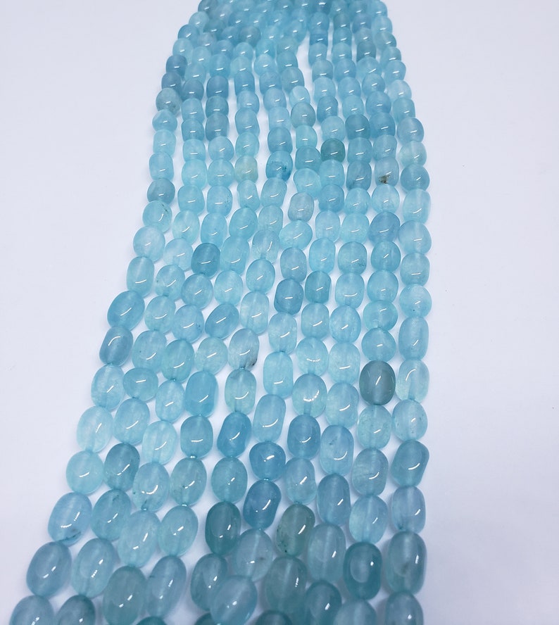 May include: Strands of aquamarine gemstone beads. The beads are oval-shaped and vary in shades of light blue. The beads are strung together, creating a cascading effect against a white background. Ideal for jewelry making.
