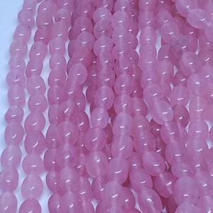 May include: Strands of polished, oval-shaped rose quartz beads. The beads are a soft pink color and have a smooth, glossy finish. The beads are strung together, creating a flowing, textured appearance. Ideal for jewelry making.