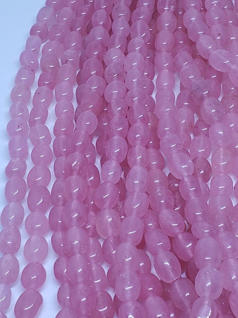 May include: Strands of polished, oval-shaped rose quartz beads. The beads are a soft pink color and have a smooth, glossy finish. The beads are strung together, creating a flowing, textured appearance. Ideal for jewelry making.