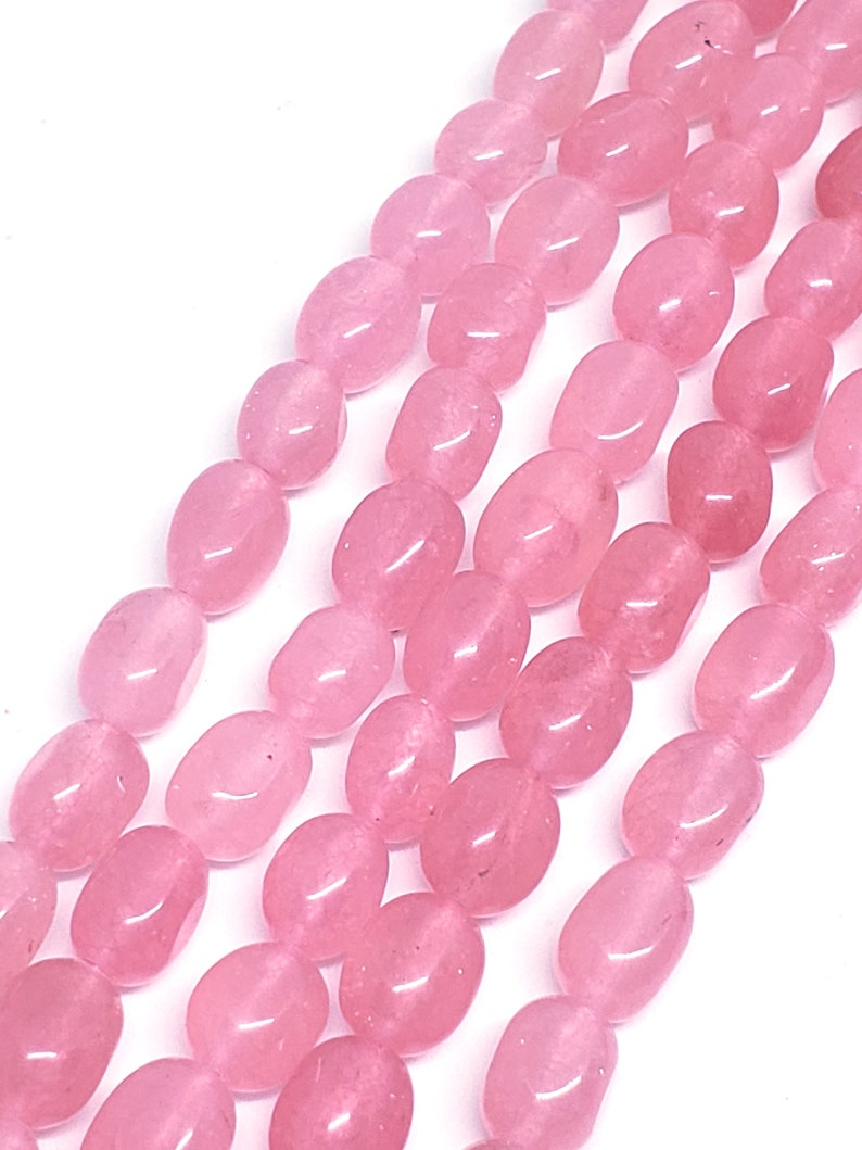 May include: Close-up of strands of polished, oval-shaped rose quartz beads. The beads are a soft pink color and have a smooth, glossy surface. The beads are strung together, creating a beautiful and natural look. Ideal for jewelry making.