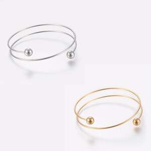 May include: Two simple, minimalist bracelets with a single ball accent on each. The bracelet on the left is silver, and the bracelet on the right is gold.