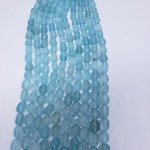 May include: Strands of aquamarine gemstone beads in varying shades of blue. The beads are oval-shaped and strung together, creating a gradient effect from light to dark. The beads are smooth and polished, reflecting light.