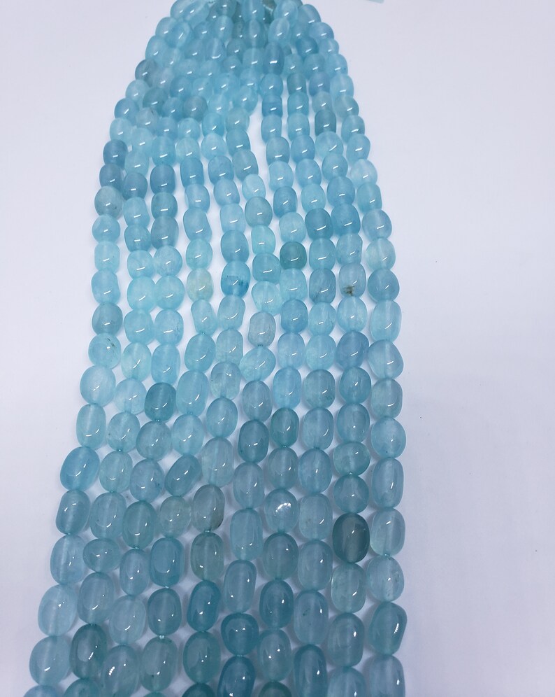 May include: Strands of aquamarine gemstone beads in varying shades of blue. The beads are oval-shaped and strung together, creating a gradient effect from light to dark. The beads are smooth and polished, reflecting light.