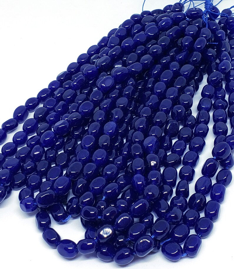 May include: A collection of deep blue, oval-shaped beads strung together. The beads have a glossy finish and are tightly packed, creating a dense, textured appearance. The beads are on a white background.