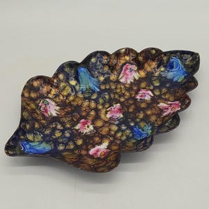 Puede incluir: Un plato decorativo en forma de hoja con una superficie texturizada. El plato presenta un fondo marrón oscuro y dorado con acentos de diseños florales azules y rosas. El plato es probablemente de cerámica o vidrio.