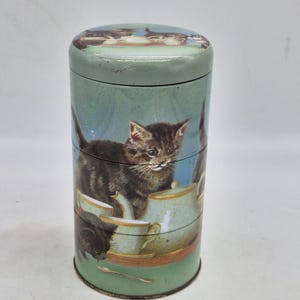Peut inclure: Une boîte de rangement cylindrique en métal sur fond vert clair, ornée d'illustrations de chatons tigrés sombres interagissant avec un service à thé. Le couvercle et le corps présentent plusieurs chatons, théières et tasses, avec une petite cuillère.