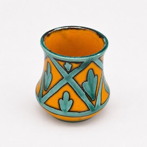 Op de afbeelding: Een kleine keramische beker met een levendige oranje basiskleur en een geometrisch rasterpatroon in donkergroenblauw. Het ontwerp bevat gestileerde bladmotieven in elk ruitvormig gedeelte en de rand is afgewerkt met een donkergroenblauwe boord.