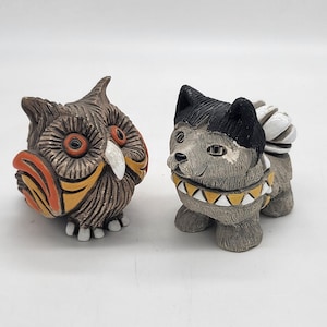 Könnte beinhalten: Zwei kleine Keramikfiguren, eine davon ein Eule mit orangefarbenen und gelben Akzenten und die andere ein Hund mit schwarzen und weißen Markierungen. Beide Figuren haben einen rustikalen, handbemalten Look.