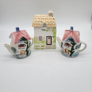Può includere: Un set di tre teiere in ceramica a forma di case. Le teiere a sinistra e a destra sono dipinte con tetti rosa, edera verde e le parole "Anna House". La teiera al centro è una casa color crema con un tetto di paglia marrone e la parola "Tea" sul davanti.