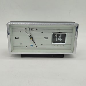 Op de afbeelding: Een rechthoekige bureauklok met een chromen frame en een witte wijzerplaat. De klok heeft zwarte wijzers, horizontale lijnen en een datumweergave met het getal 14. Boven de datum staat een diamanticoon en de tekst SHANGHAI CHINA is onderaan gedrukt.