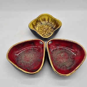 Peut inclure: Trois bols en céramique avec une finition brillante. Un bol est de forme carrée avec un intérieur jaune et noir. Deux bols sont triangulaires avec des intérieurs rouge et noir, chacun avec un petit trou sur un côté et un bord doré.
