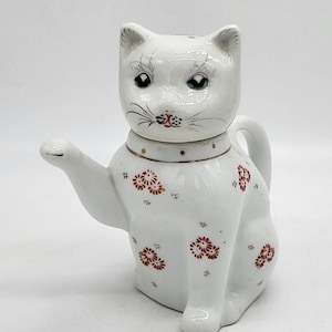 Könnte beinhalten: Eine weiße, katzenförmige Teekanne aus Keramik mit erhobener Pfote. Die Teekanne hat ein detailliertes Katzengesicht mit schwarzen und grauen Akzenten und ist mit roten Blumenmustern und kleinen grauen Strichen verziert.