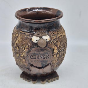 May include: A brown ceramic jar with a textured, crackled surface and a smooth, dark brown rim. The jar features a sculpted face with two white eyes, a round nose, and a bow tie. A circular plaque on the front is embossed with the word CHANGE.
