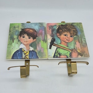 May include: Two square wall hooks featuring illustrations of children. The left hook shows a child in a blue blazer and cap. The right hook shows a child in a green shirt holding a hammer. Both hooks have gold-toned metal hardware.