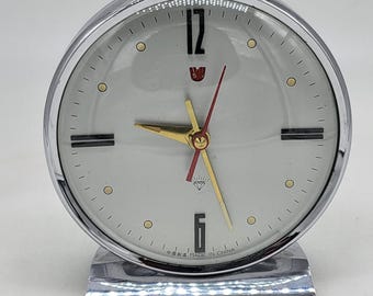 May include: A chrome-plated, round analog alarm clock with a white face. The clock features gold hands, a red second hand, and black hour markers. The text "MADE IN CHINA" is visible on the bottom. The clock sits on a rectangular base.