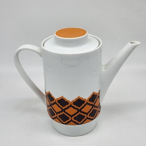 Può includere: Teiera in ceramica bianca con un motivo geometrico marrone e nero intorno alla base. La teiera ha un coperchio con la parte superiore arancione.