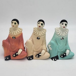 Peut inclure: Trois figurines de clown en céramique, chacune avec un costume de couleur différente. Le clown à gauche porte de l'orange, le clown du milieu porte de la crème et le clown à droite porte du bleu. Les trois clowns ont les cheveux noirs, le visage blanc, les yeux verts et des chaussures noires.