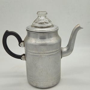 May include: Vintage aluminum coffee pot with a clear glass lid and a black handle. The pot has a spout and a cylindrical body. The lid is marked "MADE". The coffee pot is designed for stovetop use.