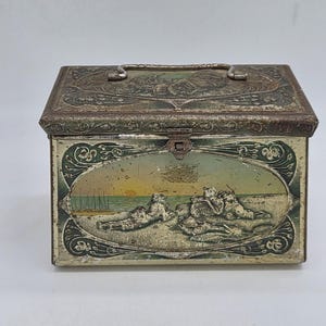 Peut inclure: Une boîte de rangement rectangulaire en métal avec un couvercle à charnière et une poignée en métal. L'avant présente une illustration ovale de personnages se relaxant sur une plage au coucher du soleil, encadrée par des volutes vert foncé.