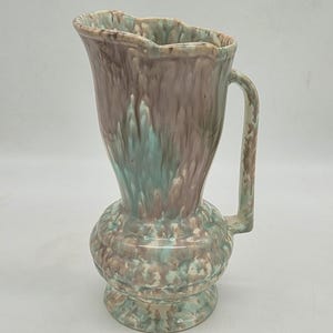 Peut inclure: Un pichet en céramique avec un émail marbré dans des tons de vieux rose, brun et vert menthe. Le récipient présente un bord large et festonné, un col étroit, une base bulbeuse au design texturé et côtelé, et une anse verticale latérale.