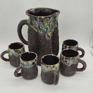 Puede incluir: Un juego de jarra y tazas de cerámica hechas a mano. La jarra y las tazas tienen un exterior marrón oscuro texturizado con acentos coloridos de verde, amarillo y azul alrededor de los bordes. El juego incluye una jarra y cinco tazas.