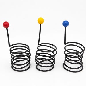 May include: Three black metal coil holders, each featuring a vertical wire extension topped with a spherical accent. The spheres are colored red, yellow, and blue respectively. These wire structures are arranged in a row against a white background.