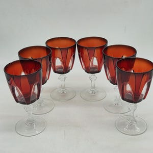 Peut inclure: Un ensemble de six verres à vin dotés de bols translucides rouge foncé avec des panneaux découpés verticaux. Chaque verre possède une tige transparente à facettes et une base circulaire, disposés en demi-cercle sur un fond neutre.