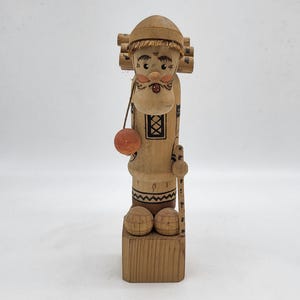 Puede incluir: Una figurita de madera de un hombre que lleva troncos en la espalda. Lleva un sombrero y tiene barba. Sostiene un palo en su mano derecha y una pequeña bola naranja en su mano izquierda. La figurita está de pie sobre una base de madera.