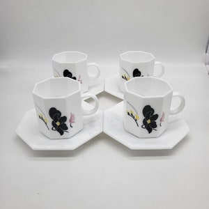 May include: Set of four white ceramic teacups and saucers with black floral designs. The cups have an octagonal shape and the saucers have a scalloped edge.