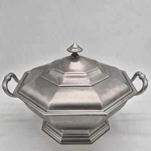 Peut inclure: Une soupière octogonale de couleur argent métallique avec un couvercle assorti et deux poignées latérales incurvées. Le couvercle présente un design à gradins surmonté d'un petit fleuron pointu. La base est également octogonale et surélevée.