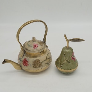 Può includere: Una teiera decorativa in ottone e un portagioie a forma di pera. La teiera ha un beccuccio e un manico curvi, con disegni floreali rossi e dorati su sfondo crema. La scatola a forma di pera è verde con accenti floreali rossi.