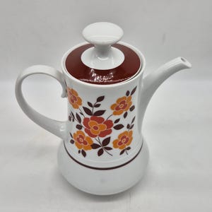 May include: A white ceramic teapot featuring a brown lid with a matching knob. The body is decorated with a floral pattern of orange and yellow flowers with dark brown leaves. It has a curved handle and a spout extending from the side.