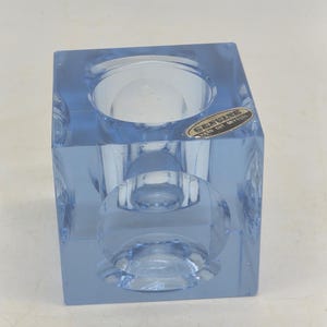 Op de afbeelding: Een lichtblauw, kubusvormig kristallen object met een cirkelvormige inkeping aan de bovenkant en zijkanten. Op de bovenzijde zit een kleine, ovale sticker met de tekst "GENUINE HAND CUT CRYSTAL" in zwarte letters op een gouden achtergrond.