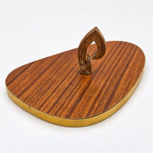 Peut inclure: Un plateau en bois en forme de goutte avec un motif de grain de bois brun riche et une finition lisse. Le plateau présente un bord doré métallique et une poignée en bois centrale, verticale et en forme de flamme fixée sur la surface.