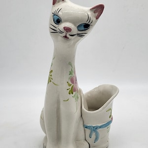 Peut inclure: Figurines de chat en céramique blanche avec des oreilles roses et des yeux bleus. Le chat est assis avec un petit pot en céramique blanche avec un ruban bleu sur le côté.
