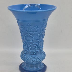 Puede incluir: Un jarrón azul claro de estilo vintage con una parte superior acampanada y un patrón floral detallado. El jarrón tiene una base bulbosa y una base azul oscuro. La altura total es de aproximadamente 20 cm.