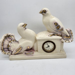 May include: A ceramic clock featuring two white doves with gold and purple accents. The clock face is round with black numerals and hands. The doves are perched on a small, brick-like structure.