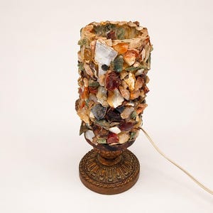 Puede incluir: Una lámpara de mesa cilíndrica con un exterior de mosaico compuesto por varios fragmentos de piedra irregulares y multicolores en tonos ámbar, marrón, blanco y gris. La lámpara descansa sobre una base circular ornamentada de color marrón oscuro.