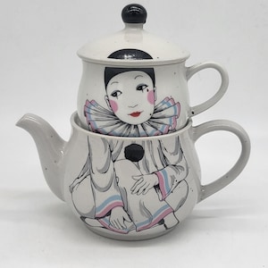 Könnte beinhalten: Zweiteiliges Keramik-Teeservice mit einem skurrilen Harlekin-Design. Die Teekanne und der Deckel sind weiß mit einer grauen Waschung und zeigen eine sitzende Harlekinfigur mit rosa, blauen und schwarzen Akzenten. Der Deckel hat einen schwarzen Knopf.