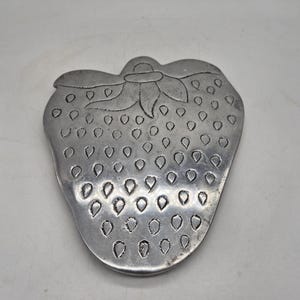 Può includere: Un sottopentola a forma di fragola, color argento. Il sottopentola presenta un design dettagliato, tra cui uno stelo e foglie nella parte superiore e piccole rientranze di semi sulla superficie. Il metallo ha un aspetto leggermente invecchiato.