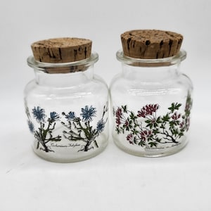 以下が含まれることがあります： コルクの蓋と花のデザインが施された透明なガラスの瓶が2つ。左側の瓶は青い花のデザインで「Cichorium Intybus」という文字が書かれており、右側の瓶はピンクの花のデザインで「Rhododendron  de  la  Montagne」という文字が書かれている。